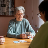 Eine altere Frau und zwei Pflegerinnen sitzen an einem Tisch und spielen Karten.