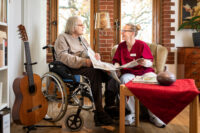 Ein Bewohner im Rollstuhl und einer Pflegerin lesen zusammen Zeitung. Beide lächeln sich an. Links an der Wand steht eine klassische Gitarre. Vor ihnen steht ein Beistelltisch mit einer roten Tischdecke.