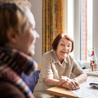 Eine ältere Frau mit dunkelroten kurzen Haaren sitzt an einem Tisch am Fenster. Sie lächelt einer Frau im Vordergrund des Bildes zu.