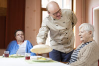 Der Koch des Hauses serviert einer Bewohnerin ihr Mittagessen. Die Bewohnerin ist begeistert von dem Essen.