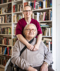 Eine Pflegerin steht hinter einem Bewohner, der im Rollstuhl sitzt. Hinter ihnen befindet sich ein Regal mit Büchern.